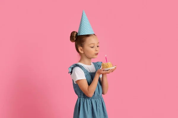 B日間の希望 誕生日ケーキの上にキャンドルを吹いてかわいい女の子 ピンクのスタジオの背景に立っている間 陽気な十代の女性の子供はパーティー帽子を身に着けています スペースをコピー — ストック写真