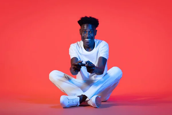 エンターテインメントとテクノロジー 幸せな陽気なハンサムな若いです黒男身に着けている白いカジュアル衣装上の床に座って ジョイスティックを保持し ネオンライトで赤の背景にビデオゲームをプレイ コピースペース — ストック写真