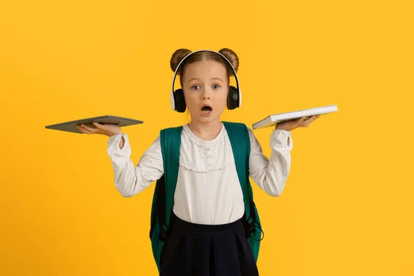 艰难的选择 震惊的小女孩拿着数字平板和纸质书的画像 小女孩在教科书和现代技术之间进行比较和选择 与黄色背景隔离 — 图库照片