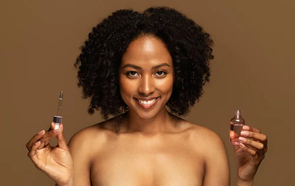 快乐迷人的 半裸的年轻黑人女子拿着装有美容品的瓶子 涂上滋养的油 与棕色背景隔离 皮肤护理常规 面部化妆品 — 图库照片