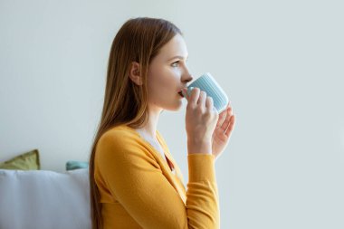 Günaydın İçecek. Sarışın genç bayan sıcak kahve içiyor, fincanla poz veriyor ve zevkini çıkarıyor, modern yatak odasında oturuyor. Yan Görünüm, Portre Çekimi