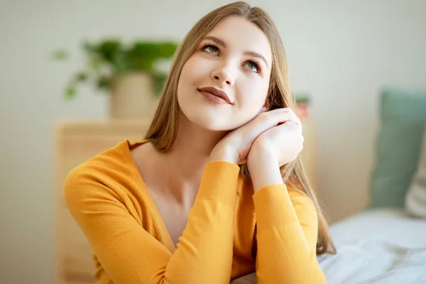 stock image Teen Dreams. Dreamy Young Blonde Lady Posing Holding Hands Near Face And Looking Up At Cozy Bedroom Interior, Sitting Near Bed Thinking About Pleasant Future. Portrait Shot