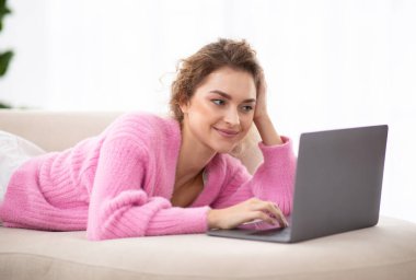 Evdeki koltukta dinlenirken dizüstü bilgisayarda internette gülümseyen genç bir kadın, bilgisayardan alışveriş yapan, cihazlara bakan ve dokunmatik dokunmatik bir dokunmatik kamera kullanan mutlu, güzel bir bayan.