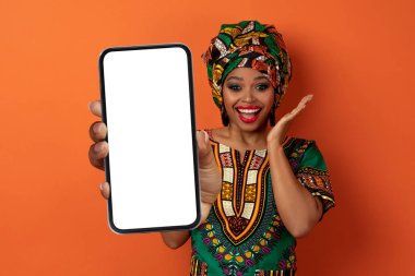 Chatbot, Al. Şaşırtıcı duygusal olarak mutlu, güzel, Afrikalı parlak bir kostüm giyen, beyaz ekranlı, hareketli, turuncu arka planda izole edilmiş, akıllı bir telefon gösteren siyah bir kadın.