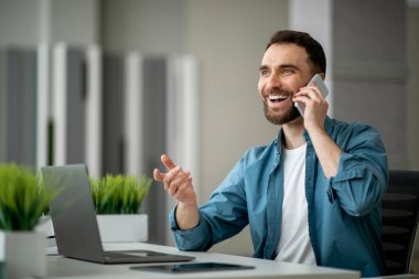 Modern ofiste çalışırken cep telefonuyla konuşan neşeli genç adam, mutlu bir iş adamı telefon görüşmesi yapıyor, keyifli cep telefonu konuşmasının tadını çıkarıyor, boşluğu kopyalıyor.