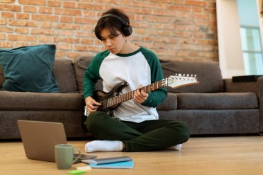 Müzikal Çevrimiçi Boş Zaman. Asyalı genç adam internetten gitar çalmayı öğreniyor. Bilgisayarı uzaktan kontrol ediyor, kulaklıkları ve ev içi bilgisayarın önünde elektrikli aletlerle oturuyor.