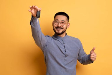 Araba kiralama. Neşeli Asyalı Adam Anahtar Tutuyor ve Sarı Stüdyo Arkaplan 'a poz verirken Başparmağını kaldırıyor, Mutlu Milenyum Erkek Kiralama Şirketi veya Yeni Otomobil Önerileri Kutlaması