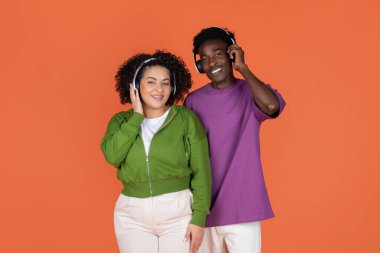 Mutlu, çoklu etnik çeşitlilikte genç bir çift Afro-Amerikan erkeği ve İspanyol bayan turuncu arka planda müzik dinliyorlar, yepyeni kablosuz kulaklıklar kullanıyorlar, kameraya gülümsüyorlar.