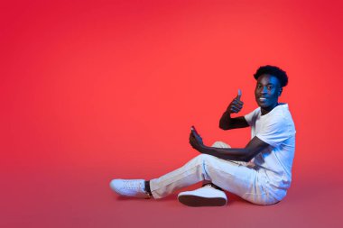 Güzel mobil uygulama. Mutlu, rahat, beyaz kıyafetli genç adam elinde akıllı telefonuyla yerde oturuyor ve baş parmağını kaldırıyor. Kırmızı arka planda, neon ışıkta izole edilmiş.