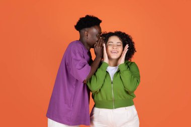 Yakışıklı Afro-Amerikan erkeği kız arkadaşının kulağına dedikodular fısıldıyor, İspanyol kadını heyecanlandırıyor, vücut ölçüleri, turuncu stüdyo geçmişi, kopyalama alanı.