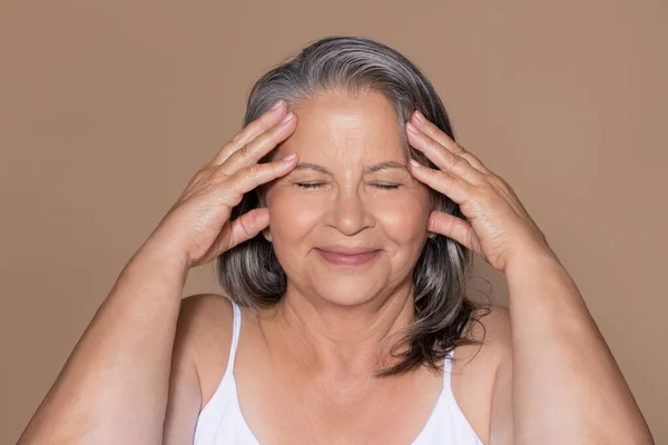 心满意足的欧洲裔老年灰发女子 闭眼按摩脸 享受提升 与米色工作室背景隔离 皮肤护理 美容程序 家庭抗衰老治疗 — 图库照片