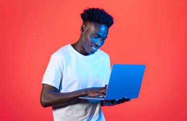 Serbest çalışma, uzaktan çalışma konsepti. Beyaz tişörtlü mutlu Afro-Amerikalı adam bilgisayarında klavyesini yazıyor ve gülümsüyor, çevrimiçi çalışıyor, kırmızı arka planda izole edilmiş, neon ışıklı, kopyalama alanı