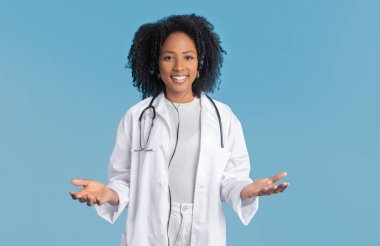 Beyaz önlüklü, mutlu, genç, Afro-Amerikan kıvırcık kadın doktor terapist mavi arka planda, stüdyoda boş bir yer tutuyor. Seçenek ilaç servisi, sağlık hizmetleri, iş