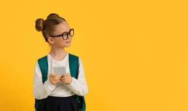 Eğitim Uygulaması. Akıllı telefon tutan ve fotokopi alanına yan bakan, elinde cep telefonu olan, sarı arka planda, Panorama 'da tek başına duran, gülümseyen küçük tatlı bir kız öğrenci.