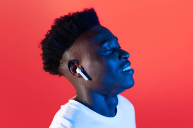 Heyecanlı, beyaz tişörtlü, yeni kablosuz kulaklıklarının keyfini çıkaran, kapalı gözlerle müzik dinleyen, gülümseyen, neon ışıklı kırmızı arka planlı mutlu siyahi adamın yakın çekimi.