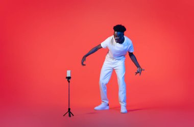 Bloglama, video bloglama konsepti. Karizmatik, havalı, şık, yakışıklı, Afro-Amerikalı bir adam tripodun önünde kırmızı arka planda, tam uzunlukta, kopyalama alanı üzerinde şarkı söylüyor.