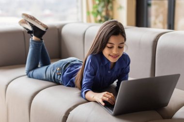 Çevrimiçi Eğlence. Gülümseyen Orta Doğulu genç kız dizüstü bilgisayar kullanıyor, oyunlar oynuyor ve evdeki koltukta uzanan web eğlencesinin tadını çıkarıyor. Dijital Hafta Sonu Boş Konsepti