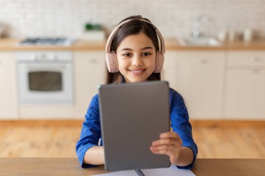 Web Eğitim Konsepti. Dijital tablet tutan neşeli liseli kız eğitim sitesini kullanıyor, kulaklık takıyor, evdeki masada oturuyor. Evinde mutlu bir çocuk portresi