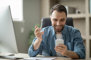 Gülümseyen yakışıklı genç işadamı modern ofiste çalışırken iş telefonu uygulamasını kontrol ediyor, mutlu milenyum erkek girişimcisi çalışma masasında oturuyor ve akıllı telefon kullanıyor, kopyalama alanı kullanıyor.