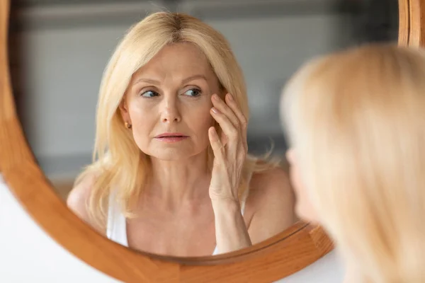 Skin aging concept. Beautiful aged woman looking at mirror at her wrinkles near eyes, checking face, noticing age changes during daily beauty routine at home
