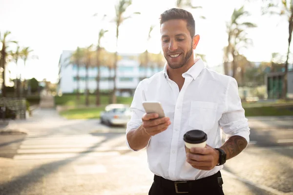 休憩中に屋外に歩いている間にスマートフォンを使ってコーヒーを飲んでいる幸せなビジネスマン 電話でインターネットを閲覧している起業家やビジネスメールに答える人 無料のスペース — ストック写真