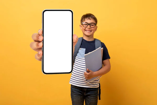 子供のためのAiチャットボット オンラインアシスタントと教育 陽気学校の白人少年は眼鏡とバックパックを着て白い空の画面で大きなスマートフォンを示しています 黄色の背景に隔離されました — ストック写真