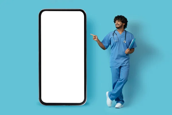 穿着医生制服的英俊的印度小伙子高兴极了 他指着装有白屏的巨大智能手机 展示了一个很好的移动应用 用于网上医生会诊 还模仿了蓝色 — 图库照片