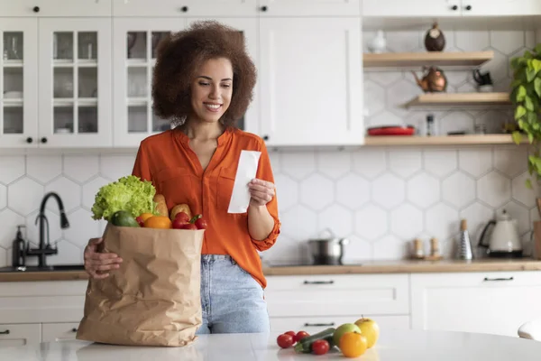 Shopping Economy. Happy Black Woman In Kitchen Checking Bill After Grocery Shopping, Smiling African American Female Standing Near Table, Counting Spends, Enjoying Affordable Prices For Food
