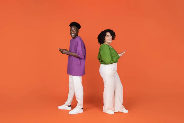 stock image E-commerce, online shopping, retail concept. Cheerful positive interracial couple in casual using cell phones isolated on orange background, paying for goods and services on Internet, full length