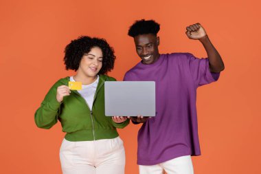 Mutlu neşeli ırklar arası aşıklar genç siyah adam ve Latin tombul kadın birlikte alışveriş yapıyor veya online bankacılık yapıyor, dizüstü bilgisayar ve banka kartı kullanıyor, izole edilmiş turuncu stüdyo geçmişi, kopyalama alanı kullanıyor.