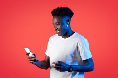 Beyaz tişörtlü mutlu yakışıklı siyah adam, kablosuz kulaklık kullanıyor ve kırmızı arka planda akıllı telefon kullanıyor, kulaklık tutuyor, internetten video izliyor, uzayı kopyalıyor.