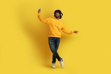 Neşeli, genç, şık, şık, yakışıklı Hintli adam. Sıradan kıyafetler giyip, sarı arka planda müzik dinlerken akıllı telefondan selfie çekiyor. Kablosuz stereo kulaklık kullanıyor.