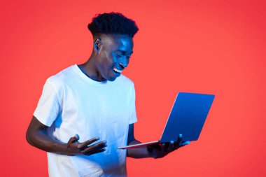 Çevrimiçi asistan, sohbet robotu, yapay zeka ve günlük yaşam. Mutlu, beyaz tişört giyen, modern bilgisayar bilgisayarı kullanan ve kırmızı arka planda neon ışıkta el kol hareketi yapan siyahi bir adam.