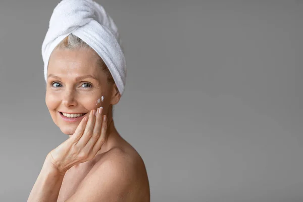 温泉与健康的概念 迷人的成熟女人在淋浴后使用面霜 头戴毛巾站在灰蒙蒙的背景上 享受美容美感和日常的面部护理 自由自在 — 图库照片