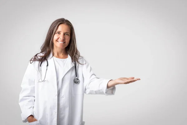 医療提供の概念 幸せなシニア女性医師は脇に指して 彼女のオープン手のひらでコピースペースを示します 新しい薬や治療を実証 光の背景にカメラで笑顔 — ストック写真