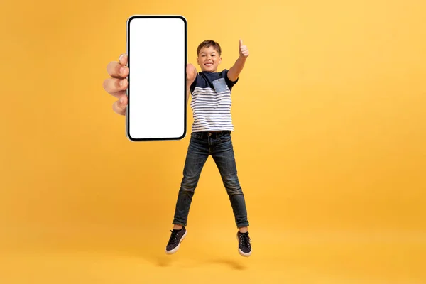 子供のためのクールなモバイルアプリ 白い空の画面と親指アップ モックアップ 黄色のスタジオの背景に隔離された コピースペースを持つ大きなスマートフォンを表示し ジャンプアップ幸せなかわいいハンサムな男の子の学校 — ストック写真