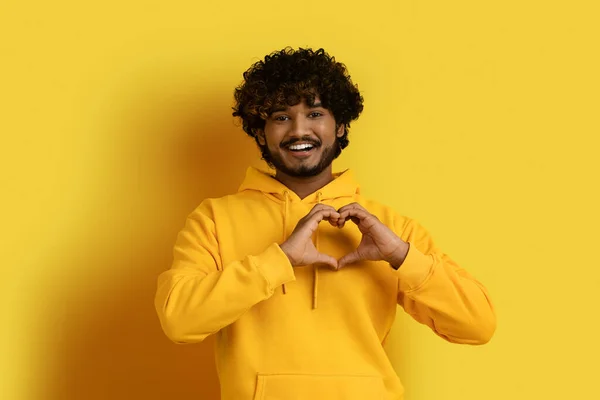 手势的概念 穿着黄色帽衫的英俊英俊的印度小伙子高兴极了 他的胸前挂着一颗心的手势 脸上挂着微笑 被黄色的背景隔开了 — 图库照片