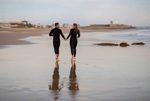 浪漫的年轻夫妇一起在沙滩上奔跑的后视镜 快乐的爱千年的男人和女人在冲浪后手牵着手在海滨散步 复制空间 — 图库照片