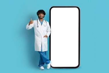 Sağlık hizmeti mobil uygulama kavramı. Beyaz önlüklü yakışıklı Hintli doktor beyaz ekranlı akıllı telefonun yanında duruyor ve baş parmağını gösteriyor, mavi arka planda izole edilmiş, maket, kopyalama alanı.
