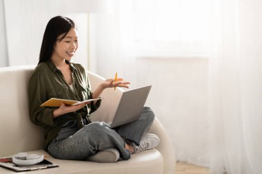 Neşeli, pozitif Asyalı bir kadın. Günlük kıyafet öğrencisi. Evinden Webinar 'a katılıyor. Kanepede oturuyor, dizüstü bilgisayar kullanıyor.