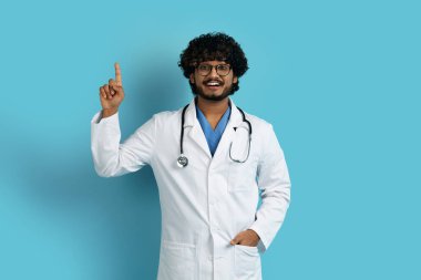 Tıp alanında çalışan genç ve neşeli Hintli bir adamın mavi stüdyo arka planında parmak kaldırması. Sağlık ve tıp alanında yenilikler