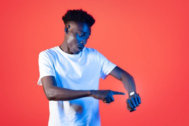 Endişeli genç, stresli, genç, yakışıklı bir adam, el bileğindeki modern ukala hareketlere bakıyor, buluşmaya ya da uçuşa geç kalıyor, parlak kırmızı arka plan, kopyalama alanı. Yaşam konseptindeki modern teknolojiler