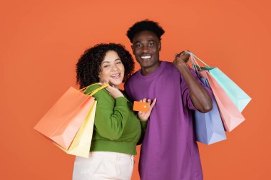 Renkli alışveriş çantaları ve turuncu stüdyo arka planında izole edilmiş banka kartı olan mutlu, çoklu etnik kökenli genç çift, siyahi adam ve İspanyol bayan temas kurmadan alışveriş öneriyor, kopyalama alanı