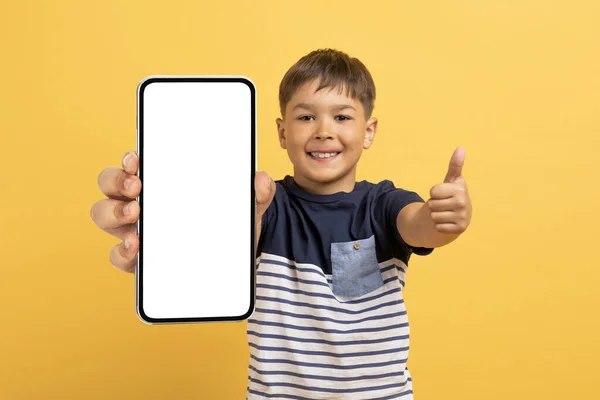 子供のためのクールなモバイルアプリ 陽気笑顔かわいいですハンサムな男の子学校ショー大きなスマートフォンで白い空の画面と親指アップ モックアップ 黄色のスタジオの背景に隔離 — ストック写真