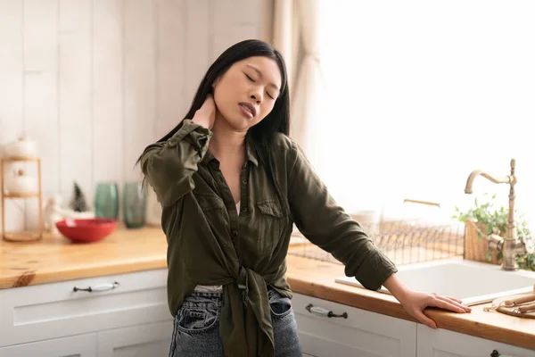 疲れ果てた若いアジアの女性は 台所に立ってカジュアルな服装の主婦を着て 机の上にもたれ 首をこすり 筋肉のひずみ コピースペースに苦しんでいる中国の女性 — ストック写真