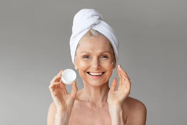 快乐的老年妇女 头上拿着毛巾 手里拿着装有保湿抗衰老霜的罐子 拍拍自己的脸 摆出一副灰色的样子 — 图库照片