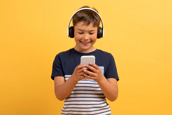 穿着休闲装 快乐可爱 英俊的学龄儿童 使用手机和无线耳机 与黄色背景隔离 开心的孩子看视频内容 在网上玩游戏 — 图库照片