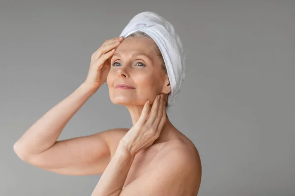 漂亮的老妇人 头戴毛巾 摸着脸 把目光移开 享受日常的面部护理 灰色背景 复制空间 温泉后的概念 — 图库照片