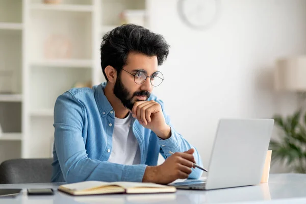 Young Indian Male Writer Working With Laptop At Home Office, Pensive Eastern Man Looking At Computer Screen, Handsome Guy Sitting At Desk And Thinking About New Book Chapter, Free Space
