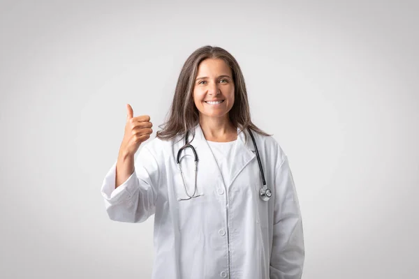 親指と笑顔を示す白いコートで興奮したシニア女性セラピスト 薬を助言 灰色の背景に隔離されたポーズ 医者の推薦 健康管理 素晴らしいサービス — ストック写真